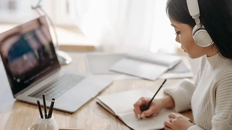woman wearing headphone writing notes in front of a laptop